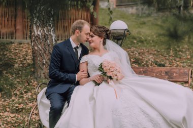 Gelin ve damat bankta oturuyor, gelin elinde bir buket çiçek tutuyor. Sahne romantik ve samimi, yeni evlilerin aşk ve mutluluğunu yansıtıyor.