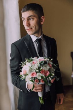 Elinde bir buket çiçek tutan takım elbiseli bir adam. Adam gülümsüyor ve pencereden dışarı bakıyor.