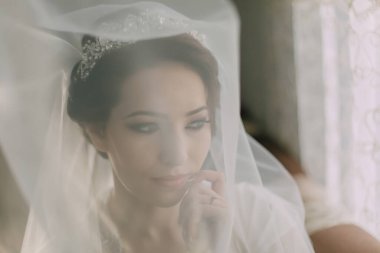 Duvak ve taç takan bir kadın kameraya bakıyor. Duvak beyaz ve yüzünü örtüyor.