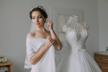 Bir kadın düğününe hazırlanıyor, duvağını takıyor ve saçını düzeltiyor. Arkasında beyaz bir elbise sergileniyor ve yanında bir manken duruyor.