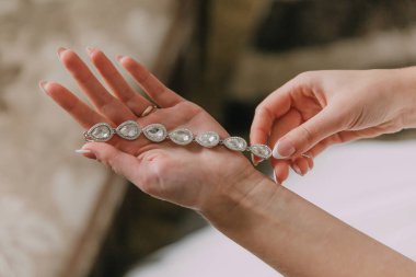 Bir kadın büyük elmas kolyeli bir kolye tutuyor. Kolye berrak camdan yapılmış ve eşsiz bir tasarımı var. Kadın beyaz bir elbise giyiyor ve kolyeye hayran.