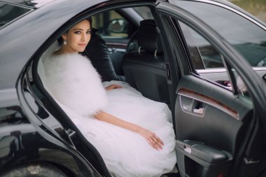 Gelinlikli bir kadın siyah bir arabanın arkasında oturuyor. Araba bir otoparka park edilmiş.