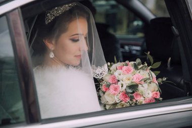 Bir gelin kucağında bir buket çiçekle arabada oturuyor. O bir peçe ve kürk manto giyiyor.