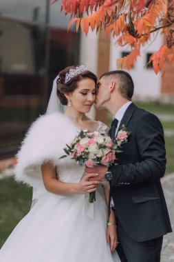 Bir gelin ve damat birbirlerinin yanaklarını öpüyor. Gelin beyaz bir elbise ve kürk şort giyiyor. Damat siyah bir takım giyiyor.