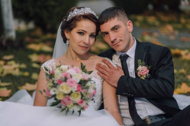 Bir gelin ve damat birlikte fotoğraf çektirmek için poz veriyorlar. Gelin beyaz bir elbise ve taç giyerken damat takım elbise ve kravat takıyor. Battaniyenin üzerinde oturuyorlar.