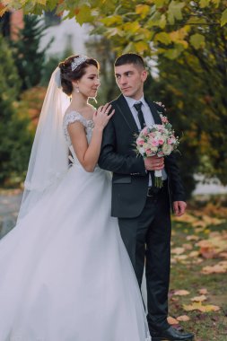 Gelin ve damat parkta, gelin beyaz bir elbise ve damat takım elbise giyerek duruyorlar. Gelin bir buket çiçek tutuyor, damat da bir vazo tutuyor.