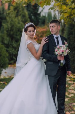 Bir gelin ve damat parkta fotoğraf için poz veriyorlar. Gelin beyaz bir elbise ve taç giyerken damat takım elbise ve kravat takıyor. Sırasıyla bir buket ve bir vazo tutuyorlar.