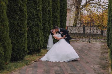 Bir gelin ve damat bahçede öpüşüyor. Gelin beyaz bir elbise giyiyor ve damat da takım elbise giyiyor.