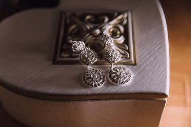 Bir çift küpe beyaz bir kutunun üzerinde duruyor. Küpeler gümüşten yapılmış ve elmas bir tasarımı var. Kutu bir masanın üzerine konur ve küpeler görüntünün odak noktasıdır.