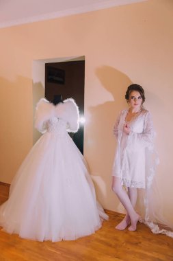 Bir kadın gelinliğin önünde duruyor, o da sergileniyor. Elbise beyaz ve uzun bir treni var. Kadın beyaz bir elbise giyiyor ve kameraya poz veriyor.