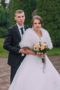 Bir gelin ve damat fotoğraf için poz veriyor. Gelin beyaz bir elbise giyiyor ve elinde bir buket çiçek tutuyor. Damat takım elbise ve kravat takıyor. Aşk ve mutluluk kavramı