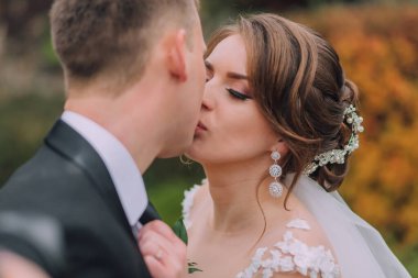 Gelin ve damat bahçede öpüşürler. Gelin beyaz bir elbise giyiyor ve damat siyah bir takım giyiyor.