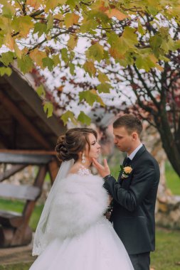 Bir gelin ve damat, yaprakları renk değiştiren bir ağacın önünde duruyorlar. Gelin beyaz kürk şort giyiyor ve damat siyah bir takım giyiyor. İkisi de gülümsüyor.