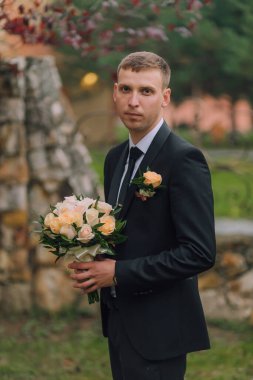 Elinde bir buket çiçek tutan takım elbiseli bir adam. Çiçekler sarı ve pembedir. Adam taş bir duvarın önünde duruyor.