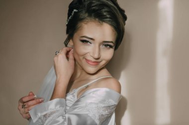 Beyaz elbiseli bir kadın gülümsüyor ve resim için poz veriyor. Kolye ve küpe takıyor.