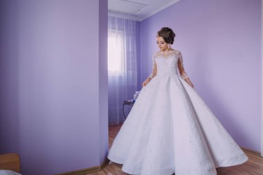 Beyaz elbiseli bir kadın mor bir duvarın önünde duruyor. Uzun, dalgalı, dantel yakalı bir elbise ve uzun bir tren giyiyor. Elbise beyaz, dantel ve nakışla ilgili küçük detaylar var.