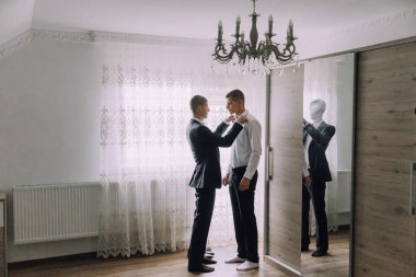 İki adam avizeli bir odada giyiniyor. Bir adam diğerine kravatını bağlamasında yardım ediyor.