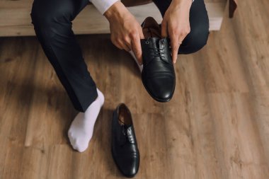 Bir adam ayakları havada ve elinde iki siyah ayakkabıyla yerde oturuyor. Ayakkabılar yan yana yere konur.