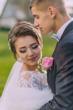 Bir gelin ve damat çimen tarlasında birbirlerini kucaklıyorlar. Gelin beyaz bir elbise giyiyor ve damat da takım elbise giyiyor.