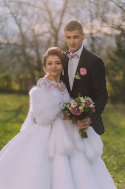 Bir gelin ve damat tarlada fotoğraf çektirmek için poz veriyorlar. Gelin beyaz bir elbise ve kürk şort giyer, damat ise takım elbise ve papyon giyer. Bir buket tutuyorlar.