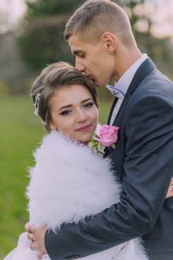 Gelin ve damat birbirlerine sarılıyorlar, gelin beyaz kürk şort giyiyor. Adam takım elbise ve kravat takıyor.