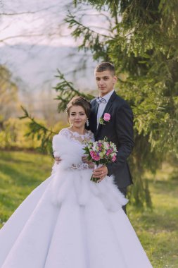 Bir gelin ve damat ormanda fotoğraf çektirmek için poz veriyorlar. Damat takım elbise giyerken gelin beyaz bir elbise ve kürk şalı giyiyor. Çiçek ve buket tutuyorlar.