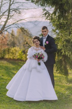 Bir gelin ve damat tarlada fotoğraf çektirmek için poz veriyorlar. Damat takım elbise giyerken gelin beyaz bir elbise ve kürk şalı giyiyor. Bir buket çiçek ve çiçek tutuyorlar.