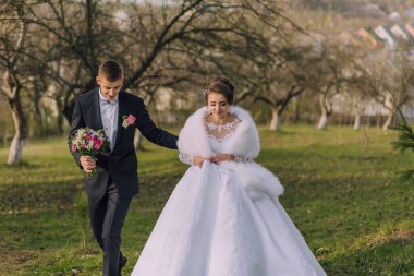 Bir gelin ve damat bir tarlada birlikte yürüyorlar. Gelin beyaz bir elbise ve kürk şort giyiyor. Damat bir buket çiçek tutuyor. Sahne romantik ve resimli.