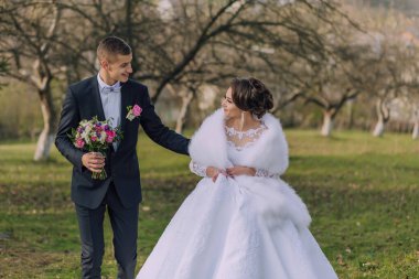 Gelin ve damat bir tarlada birlikte yürüyorlar, gelin beyaz bir elbise giyiyor ve damat takım elbise giyiyor.