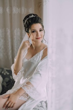 Beyaz önlüklü bir kadın pencerenin önünde oturuyor. Duvak ve taç takıyor.