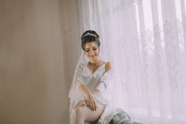 Gelinlikli bir kadın pencerenin önünde oturuyor. Duvak ve taç takıyor.