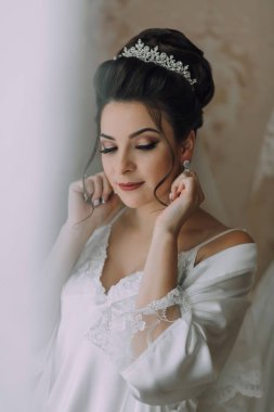 Kafasında taç olan bir kadın düğün için hazırlanıyor. Beyaz bir elbise giyiyor ve saçları topuz şeklinde.