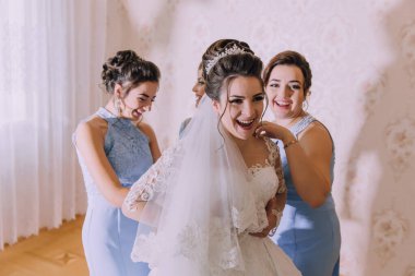 Bir gelin nedimeleriyle düğününe hazırlanıyor. Giyinmesine yardım ederken gülümsüyor ve gülüyor.