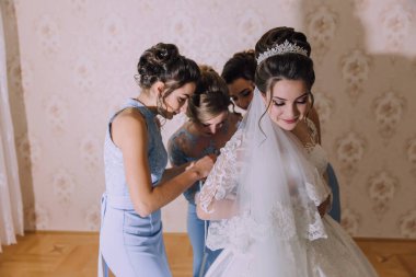 Bir gelin nedimeleriyle düğününe hazırlanıyor. Gelin beyaz bir elbise ve taç giyiyor. Nedimeler elbisesine yardım ediyor.