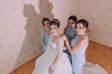 Bir gelin ve nedimeleri, çiçekli bir duvar kağıdıyla bir odada fotoğraf çektirmek için poz veriyorlar. Gelin beyaz bir elbise giyiyor ve nedimeler mavi elbiseler giyiyor. Hepsi gülümsüyor.