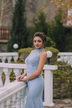 Mavi elbiseli bir kadın balkonda duruyor. Kolye ve küpe takıyor.