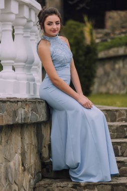 Mavi elbiseli bir kadın taş bir duvarda oturuyor. Kolye ve küpe takıyor.