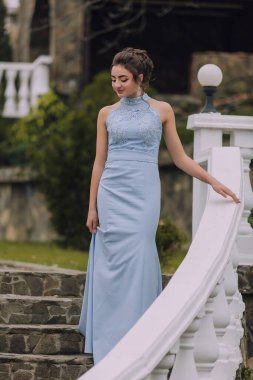 Mavi elbiseli bir kadın beyaz bir merdivende duruyor. Kolye ve küpe takıyor.