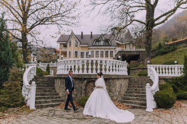 Bir gelin ve damat sarı bir evin önünde merdivenlerden iniyorlar. Gelin beyaz bir elbise giyiyor ve damat bir takım elbise giyiyor. Sahne romantik ve samimi.