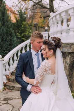 Bir gelin ve damat merdivenlerde birbirini kucaklıyor. Gelin beyaz bir elbise giyiyor ve damat bir takım elbise giyiyor. Sahne mutlu ve romantik.