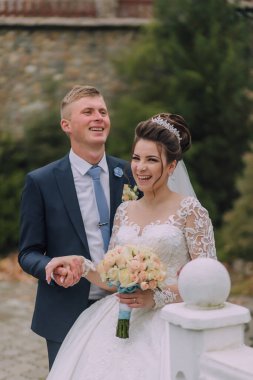 Bir gelin ve damat kameraya gülümsüyor. Gelin elinde bir buket çiçek tutuyor.