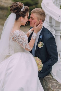 Bir gelin ve damat birbirlerini yanağından öpüyor. Gelin beyaz bir elbise giyiyor ve damat mavi bir takım giyiyor.