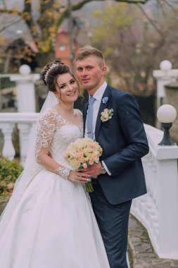 Bir gelin ve damat bir köprüde fotoğraf için poz veriyorlar. Gelin beyaz bir elbise giyiyor ve damat mavi bir takım elbise giyiyor. Bir buket çiçek ve vazo tutuyorlar.