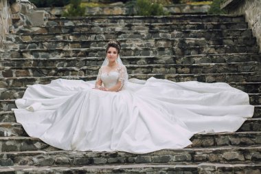 Uzun beyaz elbiseli bir kadın merdivenlerde oturuyor. Elbise uzun ve dalgalı ve kadın peçe takıyor. Zarafet ve incelik kavramı