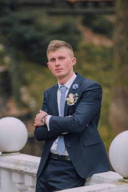 Takım elbiseli ve kravatlı bir adam köprüde kolları bağlı duruyor. Mavi bir takım elbise ve mavi bir kravat takıyor.