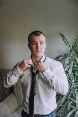 Beyaz gömlekli ve siyah kravatlı bir adam bir bitkinin önünde duruyor. Kravatını düzeltiyor ve kameraya bakıyor.