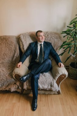 Takım elbiseli ve kravatlı bir adam kanepede oturur. O siyah bir kravat ve mavi bir takım elbise giyiyor.