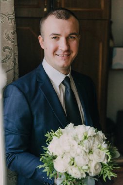 Takım elbiseli bir adam elinde bir buket beyaz çiçek tutuyor. Gülümsüyor ve kameraya bakıyor.