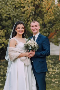 Bir gelin ve damat parkta fotoğraf için poz veriyorlar. Damat mavi bir takım giyerken gelin beyaz bir elbise ve kürk şalı giyiyor. Sırasıyla bir buket ve bir vazo tutuyorlar.