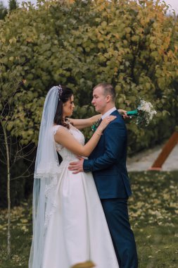 Gelin ve damat bir tarlada duruyorlar, gelin beyaz bir elbise ve damat mavi bir takım elbise giyiyor.